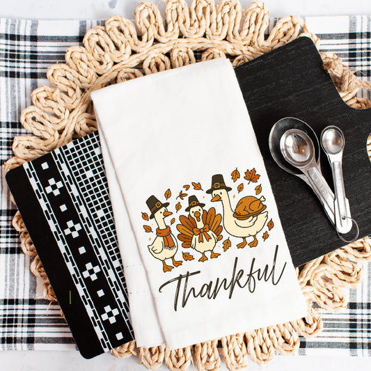 a Thanksgiving-themed kitchen towel with a turkey and autumn leaves design, along with a black cutting board, a white dish towel, and a pair of measuring spoons.