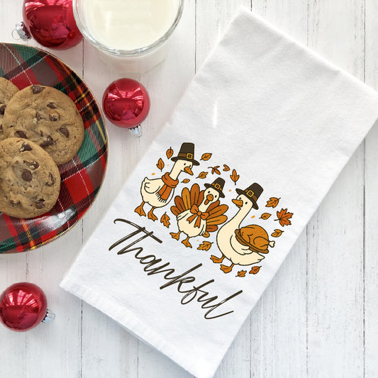A white towel with a Thanksgiving-themed design, featuring three cartoon geese wearing Thanksgiving hats and holding a turkey, along with the word "Thankful" written on it. The towel is placed on a white wooden surface, and there are a plate of cookies, a glass of milk, and ornaments, including a red ornament, arranged around it.